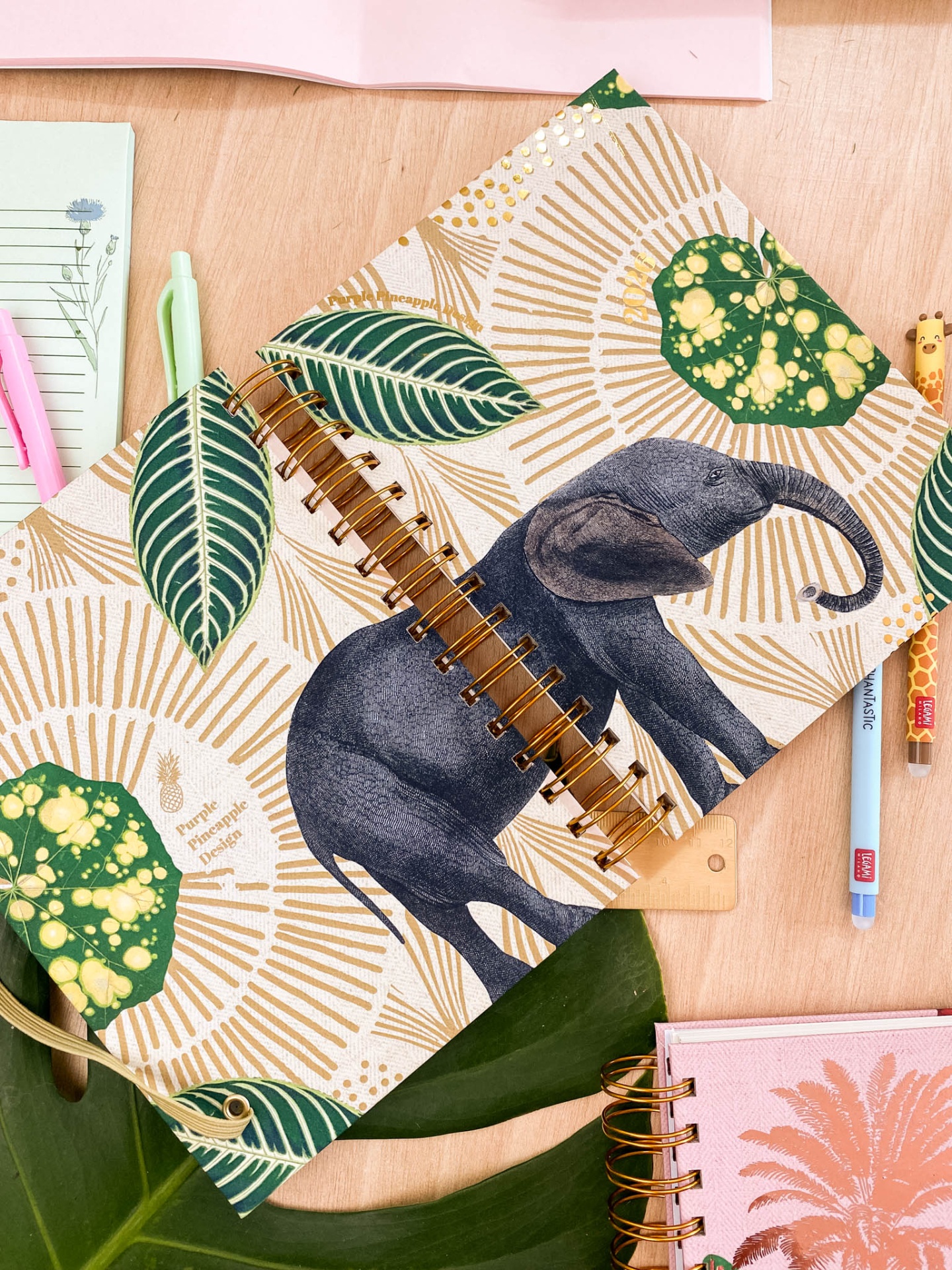Caderno espiral com elefante e folhas verde e bege sobre mesa de madeira com canetas
