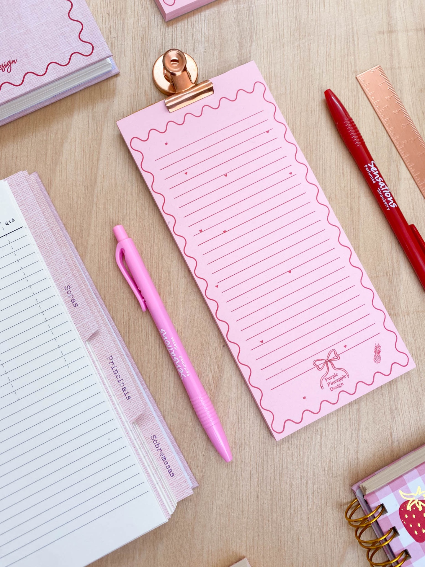 Bloco de notas rosa com canetas, régua metálica, clip e agendas numa mesa de madeira clara