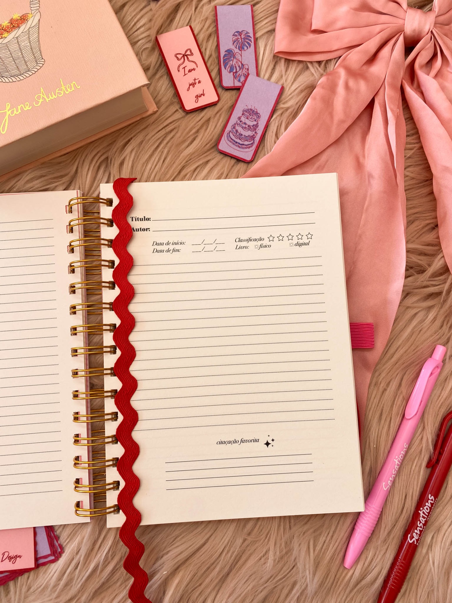Caderno rosa aberto com canetas e marcadores sobre superfície felpuda cor de bege