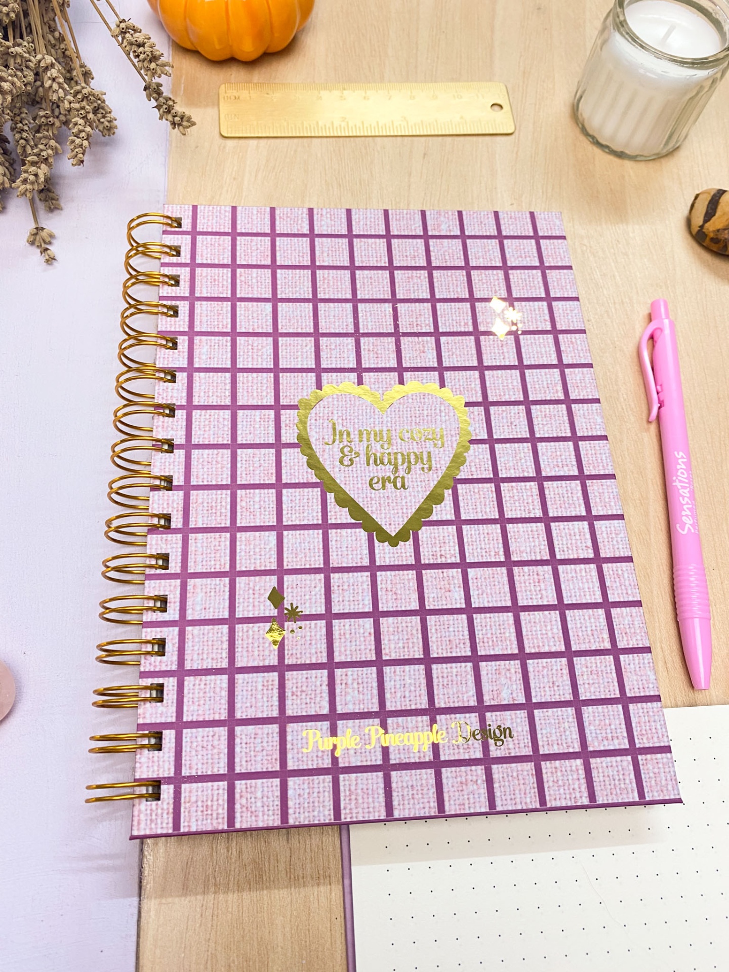 Caderno espiral rosa com padrão de quadrados, texto dourado no centro, caneta rosa e acessórios sobre superfície de madeira.