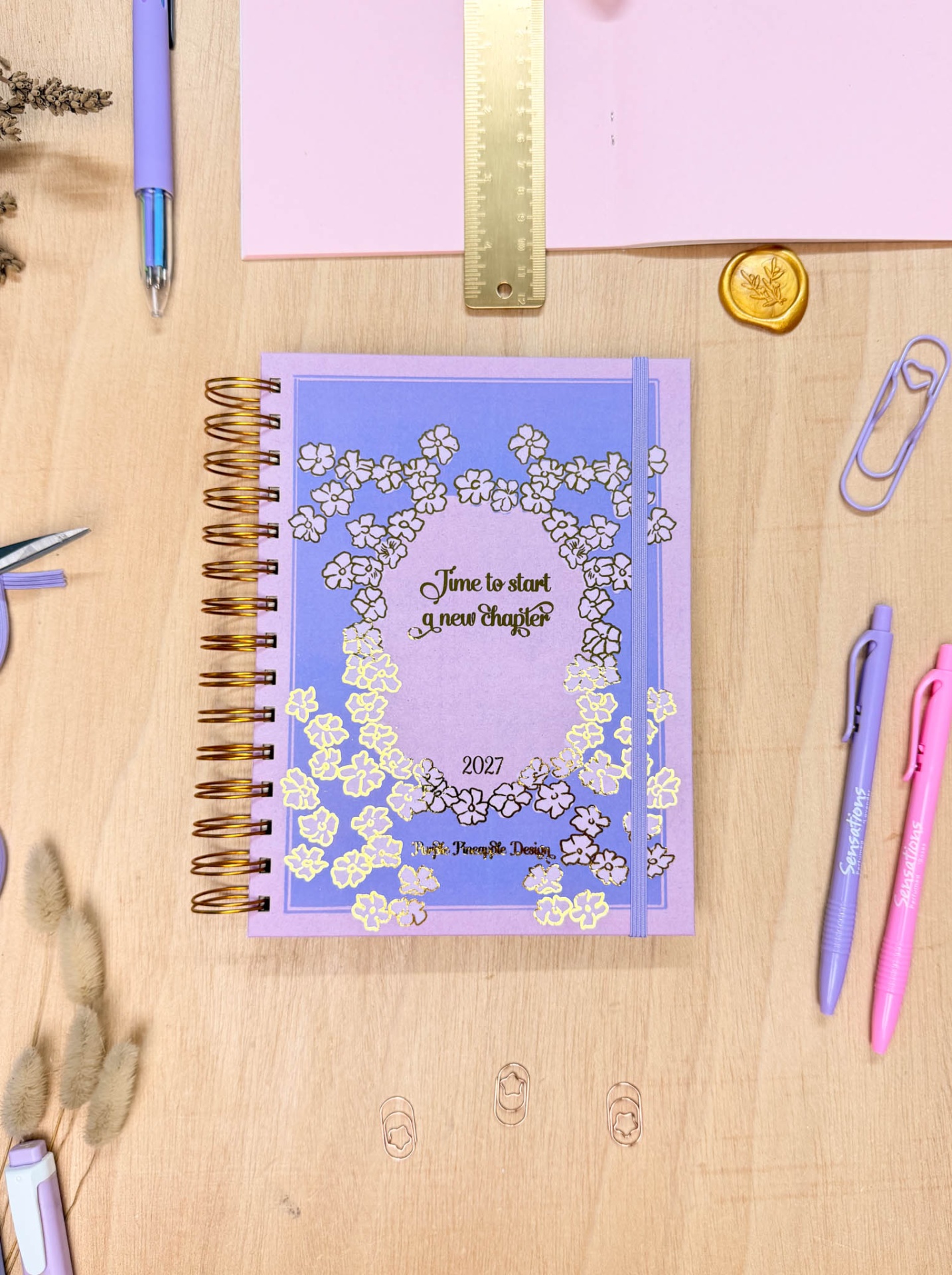 Caderno lilás espiral com design floral dourado e canetas numa mesa de madeira