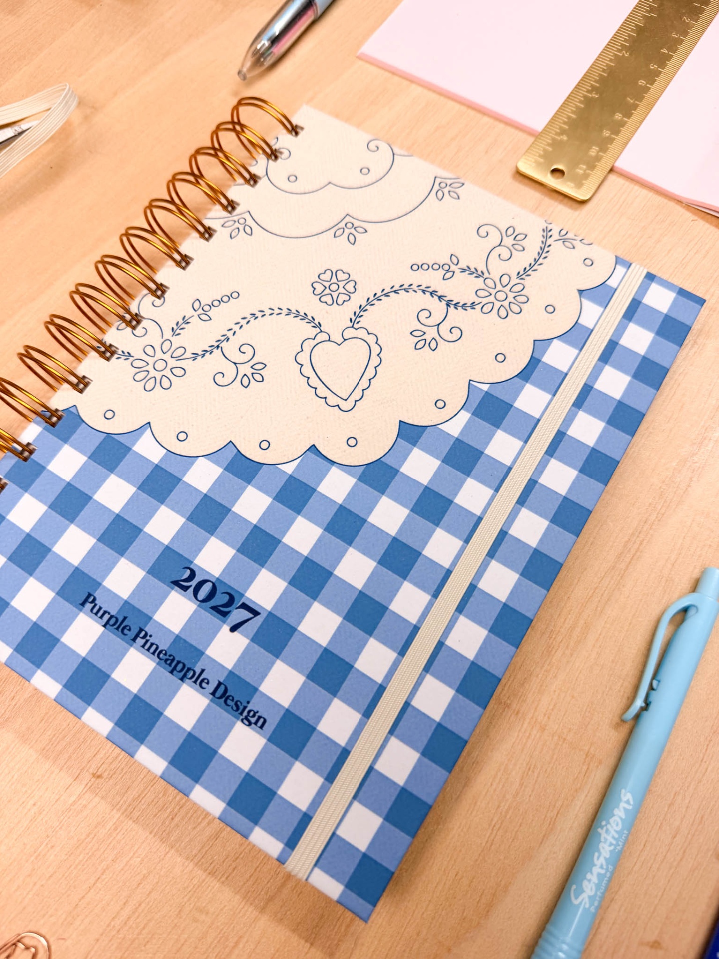 Agenda azul e branca com design de coração e flores, espiral dourada e elástico sobre mesa de madeira