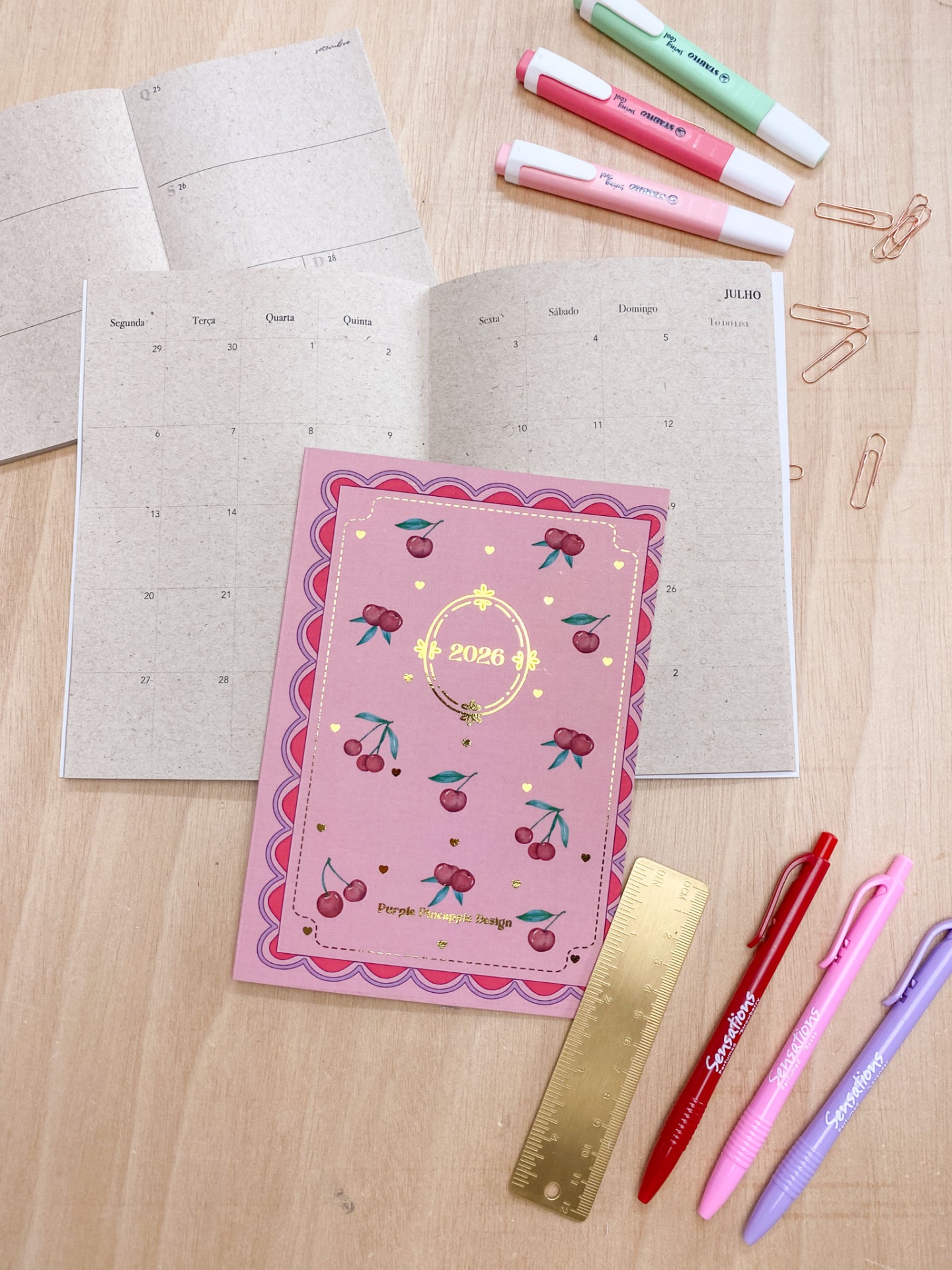 Planeador mensal 2026 com capa rosa decorada com cerejas e canetas de cores variadas numa mesa de madeira
