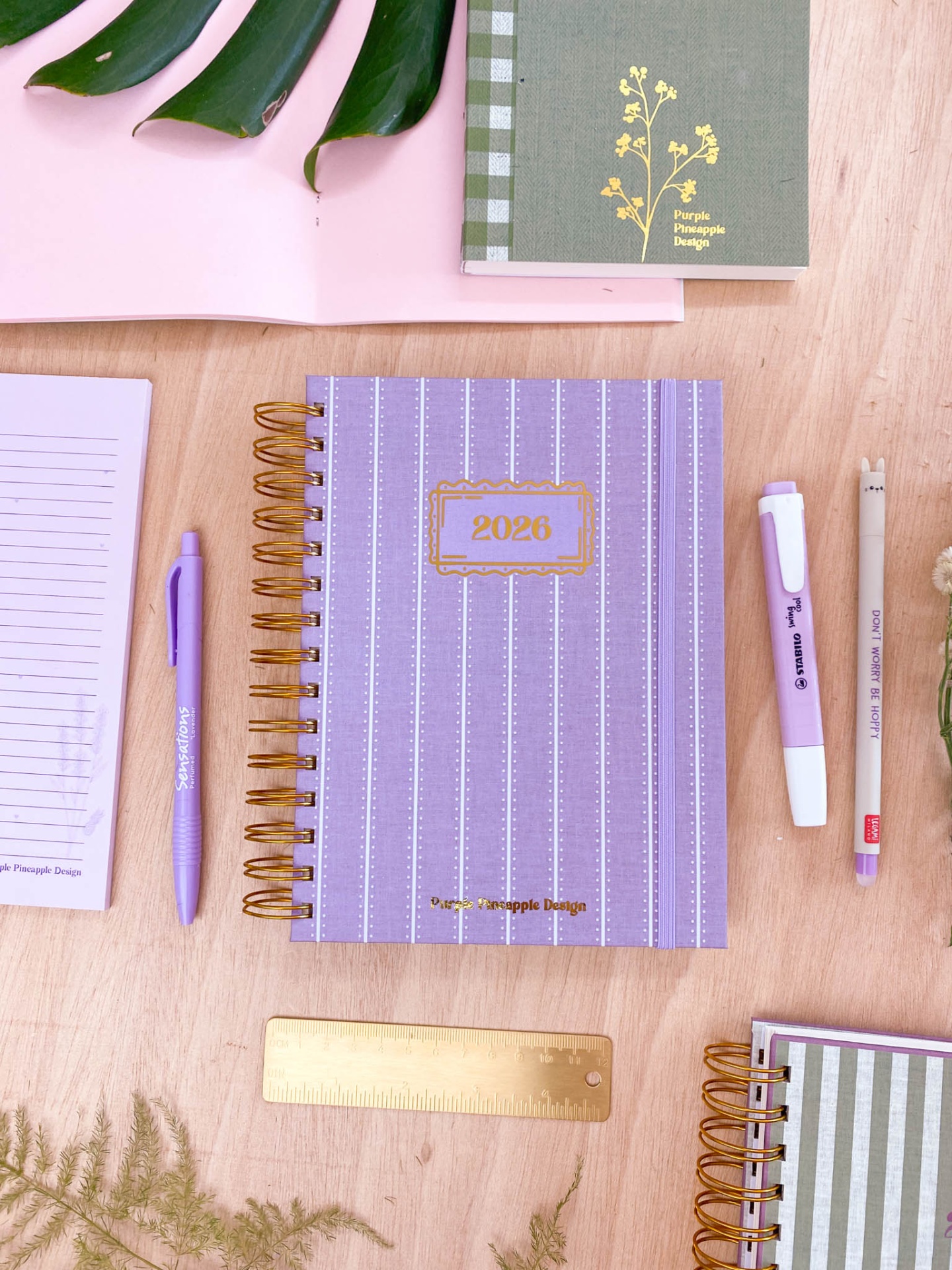 Caderno lilás 2023 com canetas, caderno verde com padrão xadrez e régua dourada sobre mesa de madeira