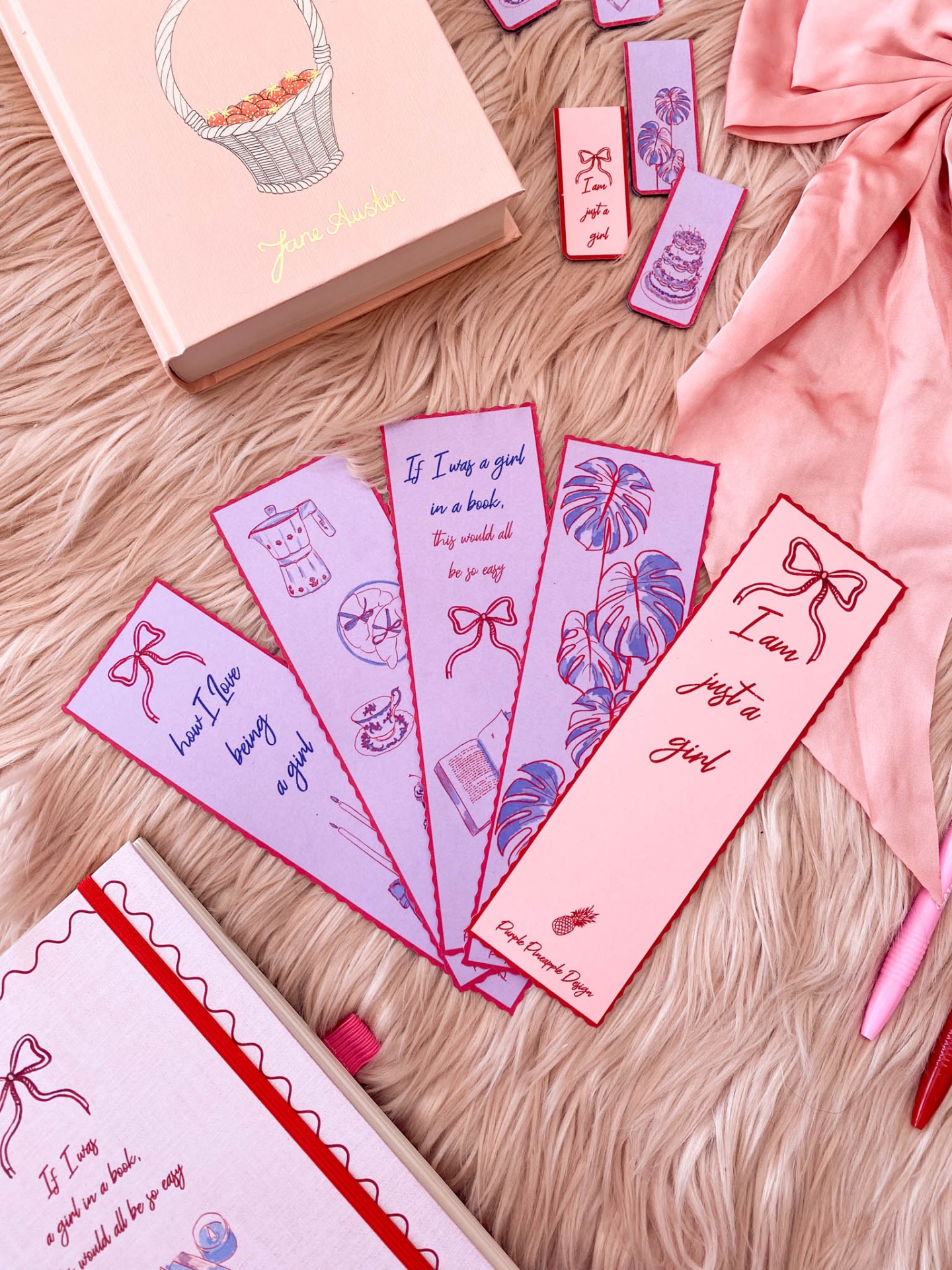 Marcadores de livros coloridos rosa e lilás com textos e desenhos, livro rosa e lenço rosa claro sobre superfície felpuda rosa.