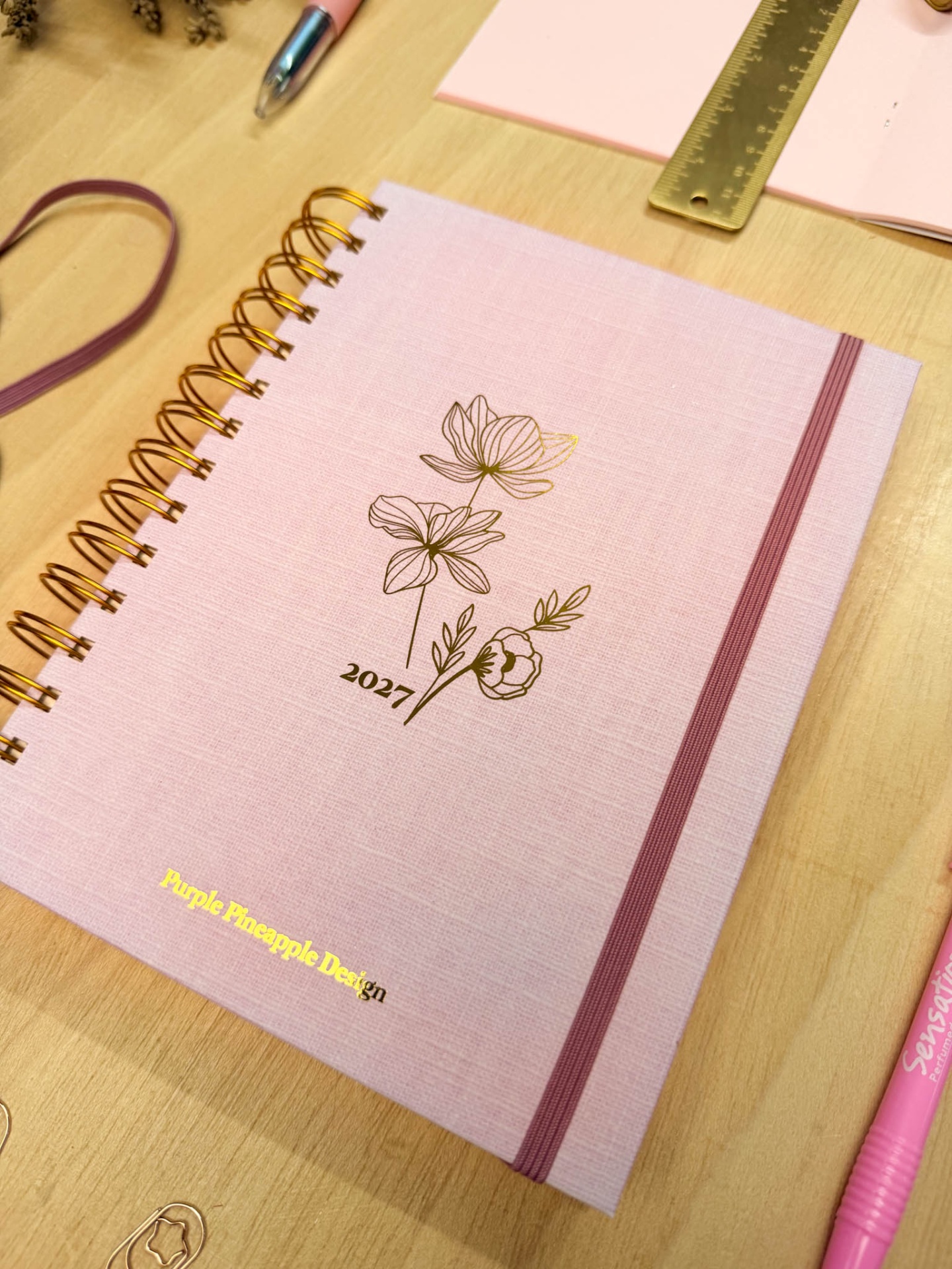 Caderno espiralado rosa claro com decoração dourada sobre mesa de madeira