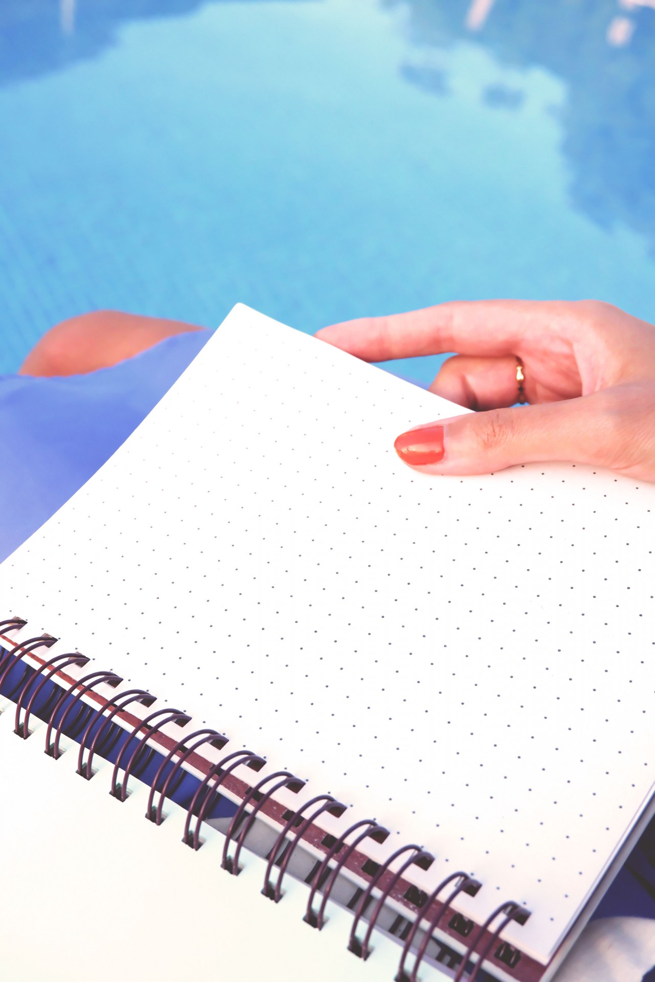 Caderno com folhas pontilhadas e espiral preto segurado por mão com unhas vermelhas próximo a uma piscina