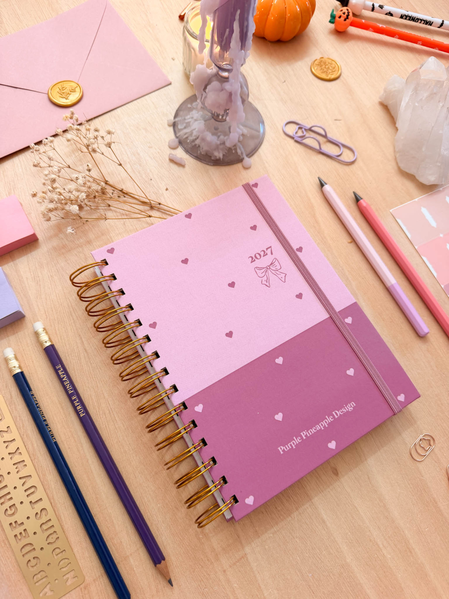 Agenda 2027 cor-de-rosa com lápis, envelope com selo dourado e vela derretida numa mesa de madeira