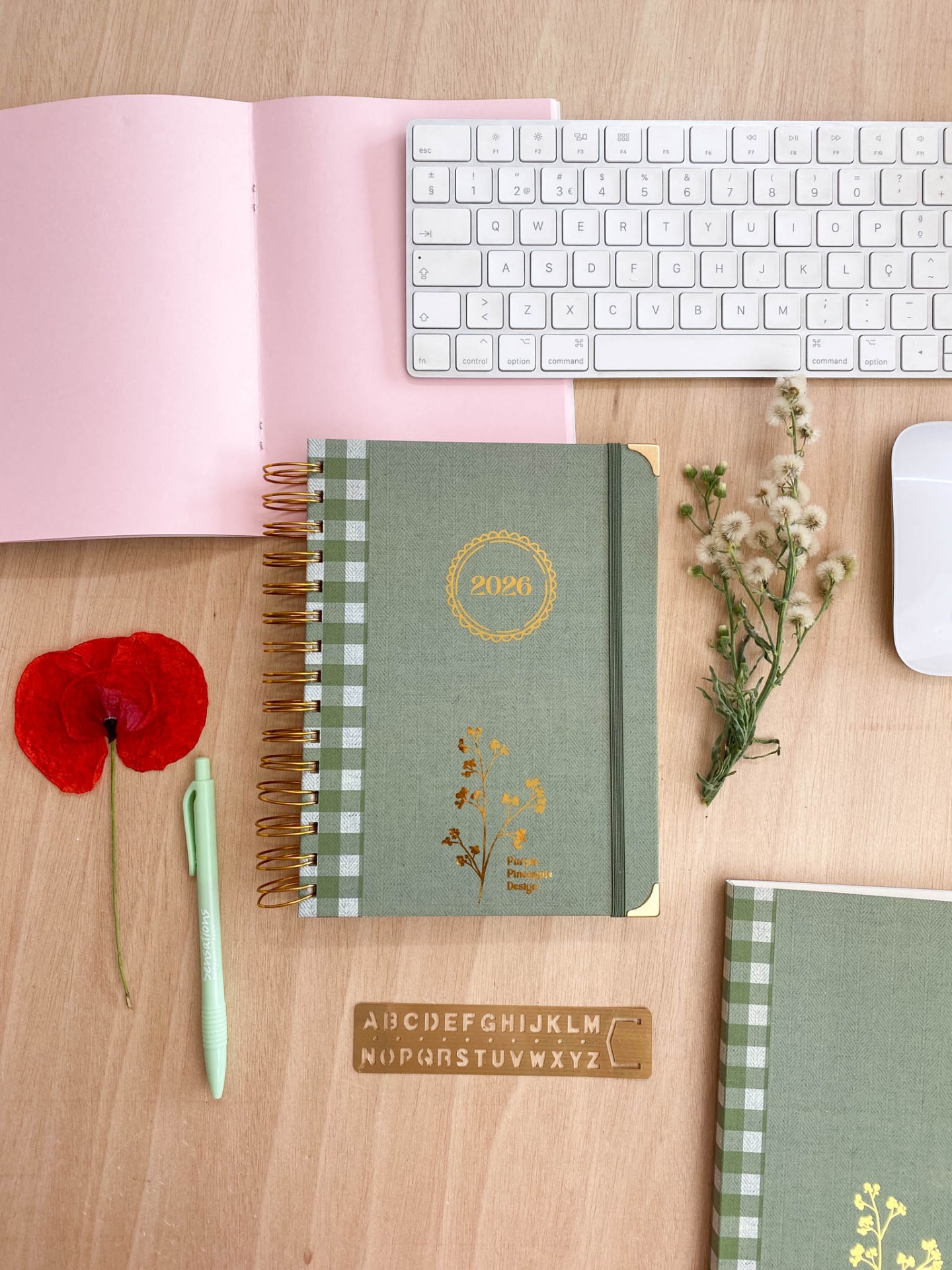 Agenda verde com espiral dourado e detalhes florais ao lado de vários objetos sobre uma mesa de madeira clara