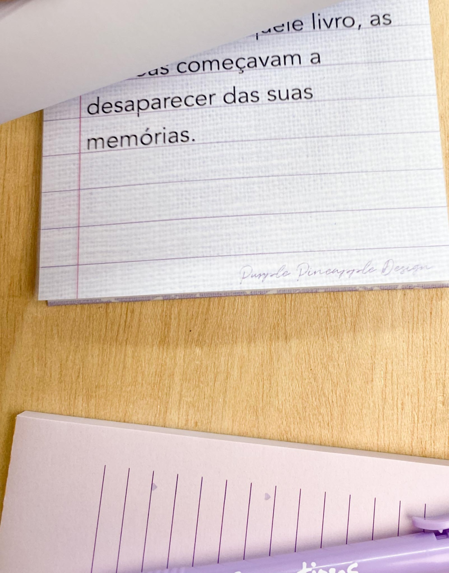 Folhas de papel branco e lilás num caderno com linhas e texto impresso, sobre mesa de madeira clara.