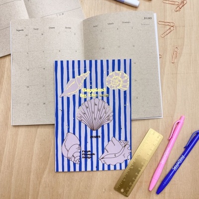 Livro com capa azul decorada com conchas e texto Amarelo ao lado de marcadores pastel numa mesa de madeira clara