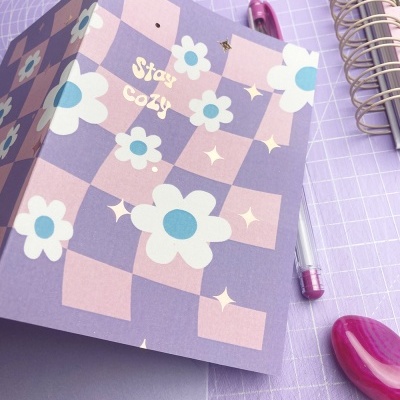 Cartão com padrão xadrez e flores, texto Stay cozy, sobre superfície lilás com caneta e caderno