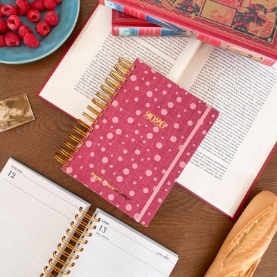 Agenda 2026 rosa com bolinhas, livros vermelhos, framboesas num prato azul e baguete numa mesa de madeira