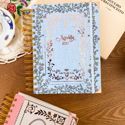 Agenda 2026 com capa floral azul e bege, mesa de madeira com livro e prato com tecido bordado