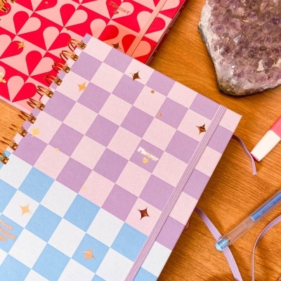 Dois cadernos coloridos espiralados com padrões distintos e elásticos, pedra ametista e canetas numa mesa de madeira