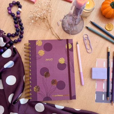 Agenda 2027 roxa com flores douradas, canetas coloridas, vela acesa, lenço roxo com bolinhas, e outros artigos de papelaria em mesa de madeira.