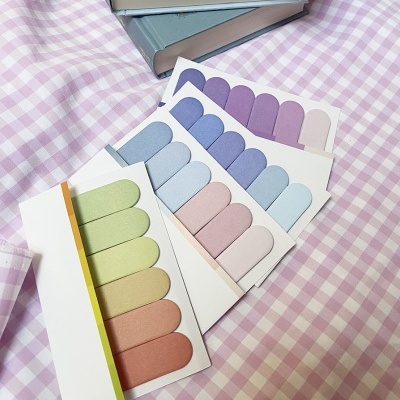 Quatro cartelas de autocolantes em tons pastel sobre tecido xadrez rosa e branco, com livros ao fundo.