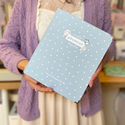 Pessoa a segurar caixa azul clara com estrelas brancas e texto, vestindo casaco roxo e blusa branca