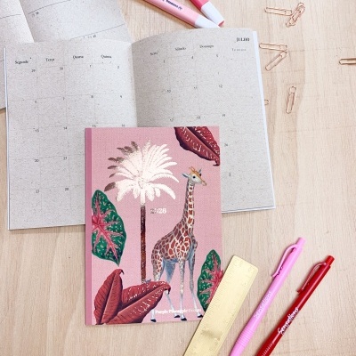 Caderno rosa com girafa e plantas, calendário aberto, canetas e marcadores rosa e vermelho, régua dourada e clips dourados