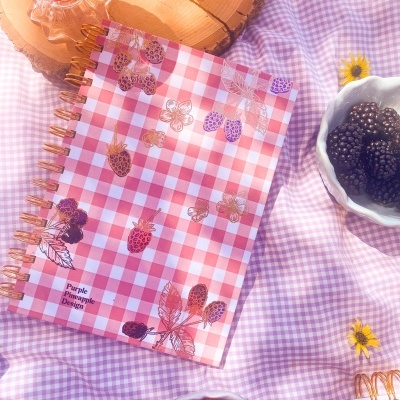 Caderno xadrez com frutas e texto Purple Pineapple Design sobre tecido lilás xadrez com tigelas de frutas e copos