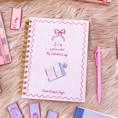 Caderno espiral branco com desenho e texto, caneta cor-de-rosa e marcadores de página em superfície felpuda