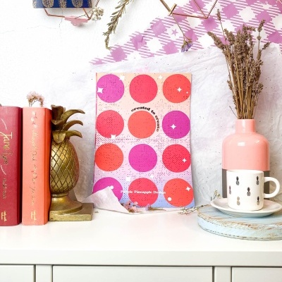 Estante decorada com livros, estatueta de abacaxi, quadro colorido, vaso com ramos secos, caneca e padrão xadrez rosa no fundo