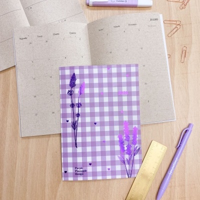 Planner aberto com capa xadrez lilás e objetos de escritório lilás e dourado sobre mesa clara