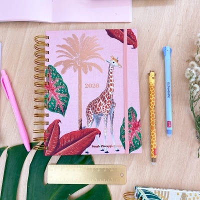 Caderno 2023 estilo tropical com girafa e folhas, canetas e lápis decorativos, régua dourada, sobre mesa de madeira