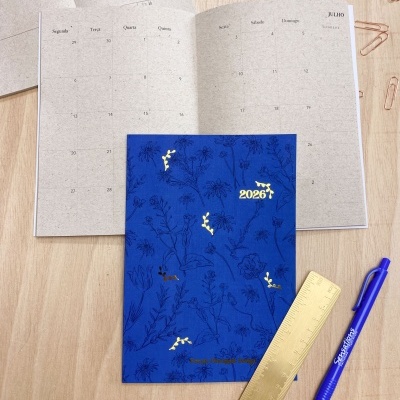 Agenda azul 2026 com ilustrações de plantas e detalhes dourados sobre mesa com régua, caneta e marcador.