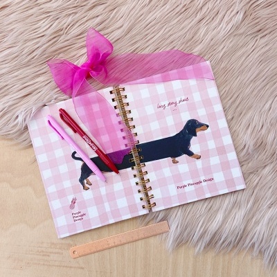 Caderno espiral com padrão xadrez rosa, cão dachshund desenhado e acessórios rosa sobre mesa