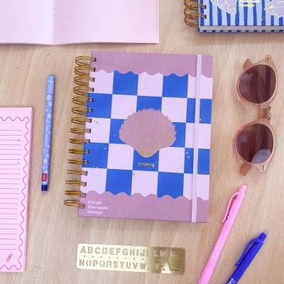 Agenda espiral com padrão xadrez azul e branco, concha dourada, canetas coloridas, régua metálica, óculos de sol e agenda azul