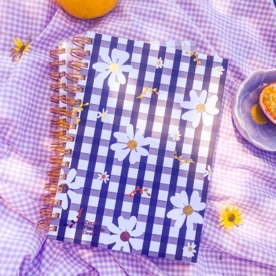 Dois cadernos espiralados com estampado xadrez azul e branco e flores, sobre tecido lilás xadrez.