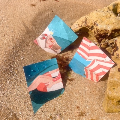 Três pedaços de papel coloridos com imagens de praia sobre areia úmida e rocha amarela