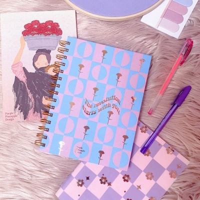 Cadernos decorados, canetas, paleta de sombras e cartão ilustrado sobre superfície peluda rosa.