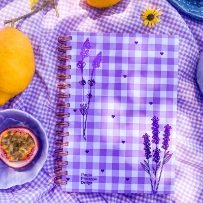 Caderno quadriculado lilás com flores desenhadas e espiral metálica sobre tecido lilás e frutas