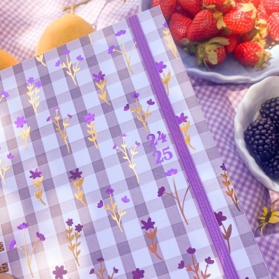 Agenda floral lilás sobre toalha xadrez com frutas em tigelas ao fundo