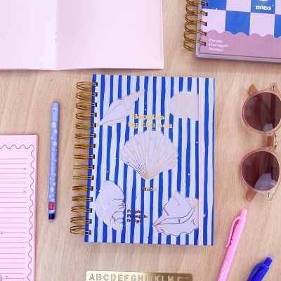 Caderno azul às riscas com conchas e texto People Pineapple Design rodeado por material de escrita e óculos de sol numa mesa