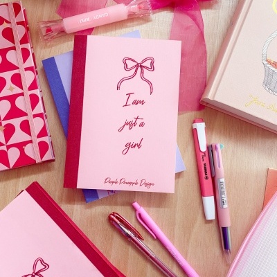 Cadernos e canetas coloridos sobre uma mesa clara, destaque para caderno rosa com texto 'I am just a girl'.