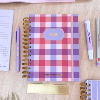 Agenda 2026 quadriculada rosa e lilás com capa espiral dourada e canetas numa mesa de madeira
