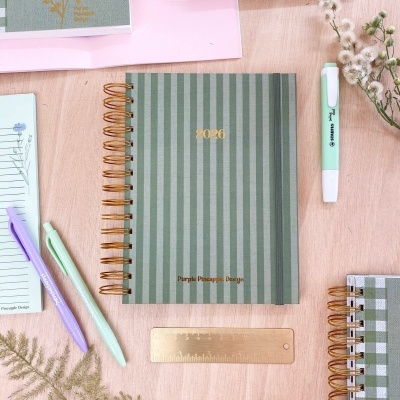 Caderno verde às riscas 2026 com canetas e régua dourada sobre mesa de madeira