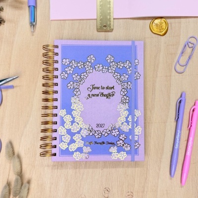 Agenda anual roxa espiralada com flores douradas na mesa ao lado de canetas e clips