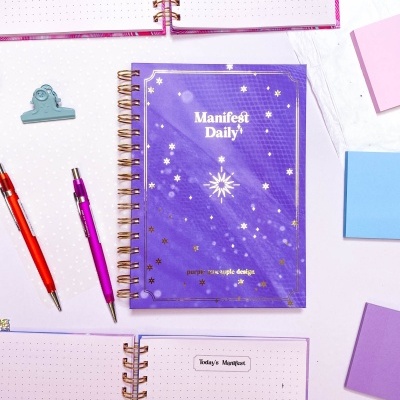 Caderno roxo Manifest Daily com estrelas douradas em fundo branco com canetas e blocos de notas coloridos