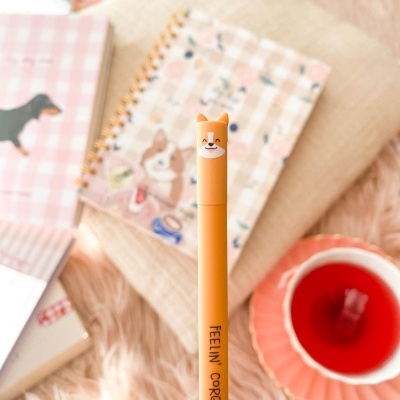 Caneta laranja com tampa em forma de cabeça de cão e texto FEELIN' GORGEOUS, com fundo desfocado de caderno xadrez e chá em xícara rosa.
