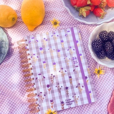 Agenda espiral com capa xadrez lilás e flores sobre tecido cor-de-rosa com frutas e flores ao redor.