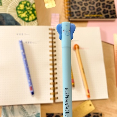 Caneta azul claro com tampa de elefante e texto ELEPHANTASTIC sobre mesa com cadernos e réguas