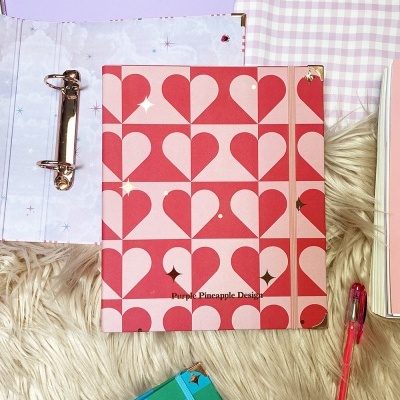 Caderno rosa e vermelho com corações e argolas metálicas sobre tapete felpudo com caneta rosa ao lado