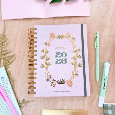Agenda 2026 com capa rosa e ilustração floral, espiral dourada e elástico cinza numa mesa de madeira com canetas e régua