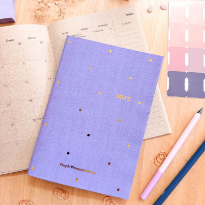 Agenda anual com capa roxa e pontos dourados sobre mesa com calendário, canetas e clips