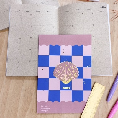 Agenda com capa xadrez azul e rosa com concha dourada e texto 2023, aberta em julho, sobre mesa com canetas, clips e régua.
