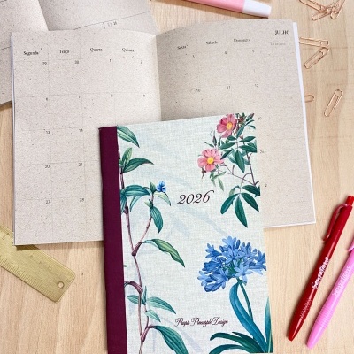 Agenda 2026 com capa floral e calendário aberto sobre mesa com material de escrita.
